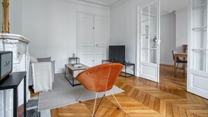 A spacious living room with a fireplace, a television, a comfortable orange chair, and a dining area with a table and chairs. - Blueground | Champ de Mars, walk to Eiffel Tower (Paris)