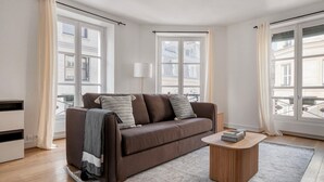 Una sala de estar moderna con un sofá marrón, una mesa de centro de madera y vistas a edificios a través de amplias ventanas.