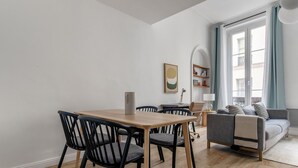 Una sala de estar moderna con zona de comedor, que incluye una mesa y sillas de madera, un sofá gris y un ventanal con cortinas.