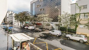 A modern building with a dark facade and multiple windows. There are parked cars and a bus stop on the street in front.