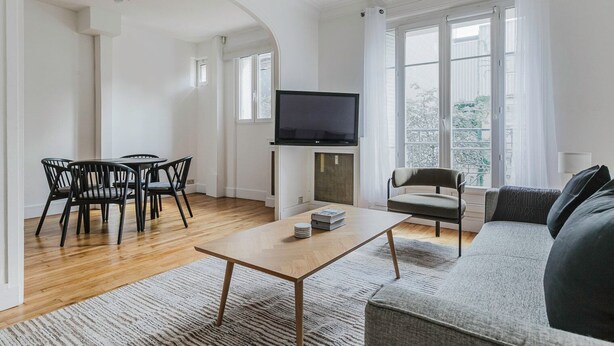 A modern living room with a wooden floor, a sofa, a TV, and a dining area with chairs.