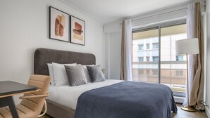 Habitación de hotel con cama, una silla, una mesa y ventana con vista a edificios.