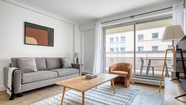 Una sala de estar moderna con un sofá gris, una mesa de centro de madera, un tapete estampado, un sillón pequeño y un ventanal con vistas a edificios.