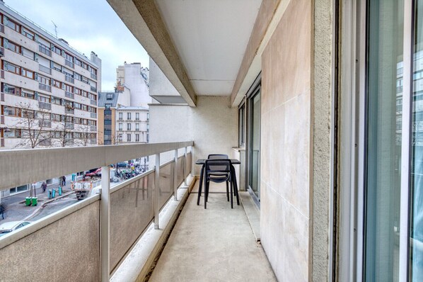 A narrow balcony with a table and chairs, overlooking a street with parked cars and buildings.