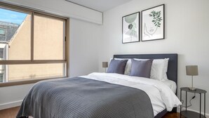 Une chambre avec une grande fenêtre, un lit avec une literie grise et blanche, deux œuvres d’art encadrées accrochées au mur et une table de chevet avec une lampe.