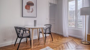 A room with a round wooden table, two black chairs, a white floor, a framed abstract painting, a window with white curtains, and a floor lamp.
