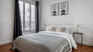 Una habitació d'hotel amb un llit gran, una finestra amb cortines, una tauleta de nit amb una làmpada i dues obres d'art penjades a la paret.