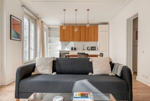 A modern living room with a dark grey sofa, two pillows, and a coffee table with books. A kitchen with wooden cabinets and a dining area is visible in the background. - Blueground | Batignolles, near park, family area (Paris)