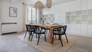 A dining area with a wooden table and black chairs, white walls, and a large window with curtains. - Blueground | Pigalle, nr restaurants & boutiques (Paris)
