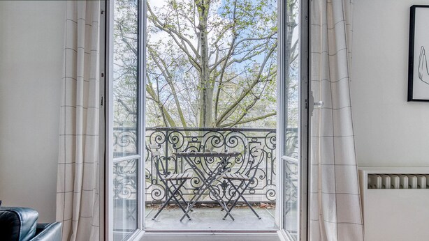 A room with a large window, a metal balcony railing, and a view of trees outside. - Blueground | Pigalle, nr restaurants & boutiques (Paris)