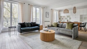 A spacious living room with a black leather sofa, a grey sofa, and a wooden coffee table. There is a dining area with a wooden table and chairs. The room has large windows with curtains and a balcony with a wrought iron railing. - Blueground | Pigalle, nr restaurants & boutiques (Paris)