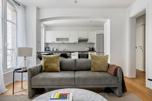Ein modernes Wohnzimmer mit einem grauen Sofa, zwei Kissen, einem kleinen Tisch mit einem Buch und einer Lampe. Im Hintergrund ist eine Küche mit weißen Schränken und Geräten zu sehen.