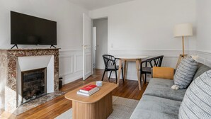 A living room with a fireplace, a flat-screen TV mounted above it, a dining table with chairs, and a sofa with cushions. - Blueground | Jardin des Plantes, nr Notre Dame (Paris)