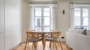 A room with a round wooden table and four chairs, a grey sofa, and a balcony with white curtains. - Blueground | Batignolles, near park & restaurants (Paris)