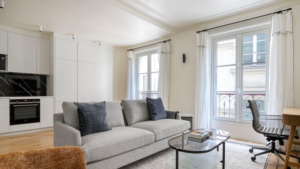 A modern living room with a grey sofa, a brown armchair, a glass coffee table, and a dining area with a chair and table. - Blueground | Batignolles, near park & restaurants (Paris)