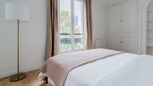 A bedroom with a bed, a floor lamp, a window with curtains, and a white wardrobe. - Blueground | Batignolles, near park & dining (Paris)