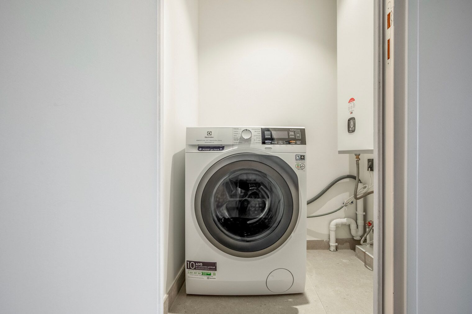 A compact, white front-loading washing machine in a narrow hallway with a visible water pipe and a small step leading to a lower level.
