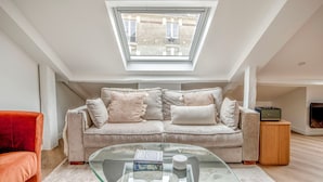 Un salon de style industriel, avec un plafond haut et une verrière, un canapé beige agrémenté de coussins, une table basse en verre et une petite table d’appoint en bois.