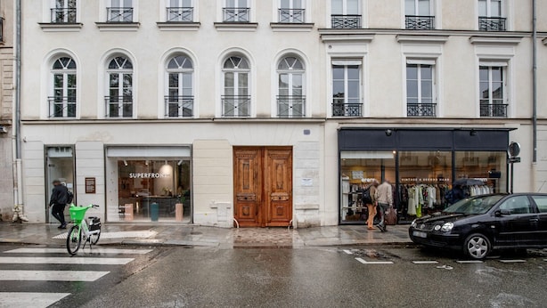 Une vue de rue avec un bâtiment doté d’une grande porte en bois et d’une devanture de magasin sur laquelle on peut lire l’inscription « SUPERFRONT ».