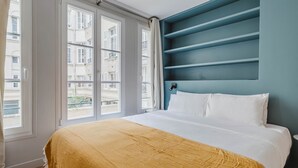 A bedroom with a bed, a window with curtains, and a blue wall with shelves.