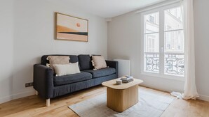 A modern living room with a dark blue sofa, a wooden coffee table, and a large window with a view of buildings.