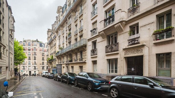A narrow street lined with multi-story buildings, parked cars, and a tree. - Blueground | Lamarck - Caulaincourt, nr dining (Paris)
