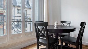 A room with a round table and four chairs, a white vase on the table, and a view of buildings through large windows. - Blueground | Lamarck - Caulaincourt, nr dining (Paris)
