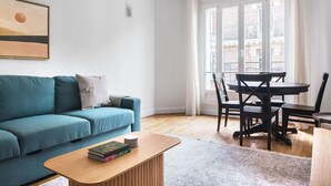 A modern living room with a teal sofa, a wooden coffee table with books, a dining table with chairs, and a large window with a view of buildings. - Blueground | Lamarck - Caulaincourt, nr dining (Paris)