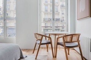 A room with a large window, two wooden chairs with cushions, a small round table, and a bed with a grey blanket. - Blueground | Le Marais-Republique, nr food & shops (Paris)