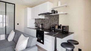 Eine moderne Küche mit weißen Schränken, einer schwarzen Arbeitsplatte und einem eingebauten Backofen. Ein graues Sofa mit einem weißen Kissen und ein schwarzer Hocker sind ebenfalls vorhanden.