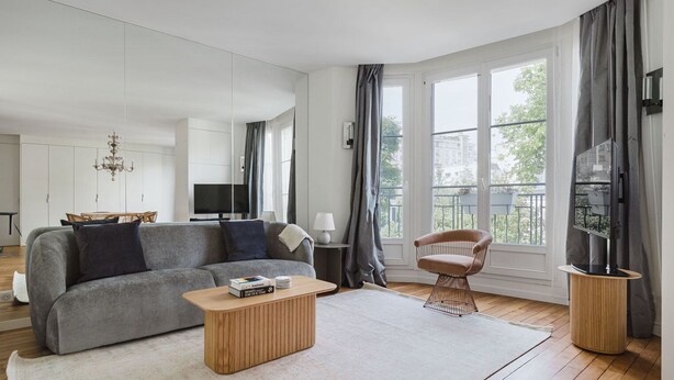 Una sala de estar moderna con un sofá gris, una mesa de centro de madera y una silla. Se ve un televisor sobre un mueble, un candelabro y un balcón a través de las ventanas.