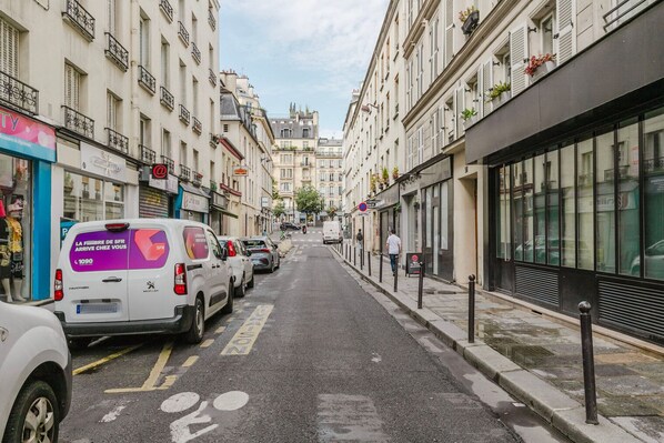 A narrow urban street lined with multi-story buildings, parked cars, and a pedestrian.