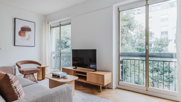 Un salon moderne avec un canapé, un fauteuil, un téléviseur posé sur un meuble en bois, et de grandes fenêtres donnant sur un balcon et un espace verdoyant.