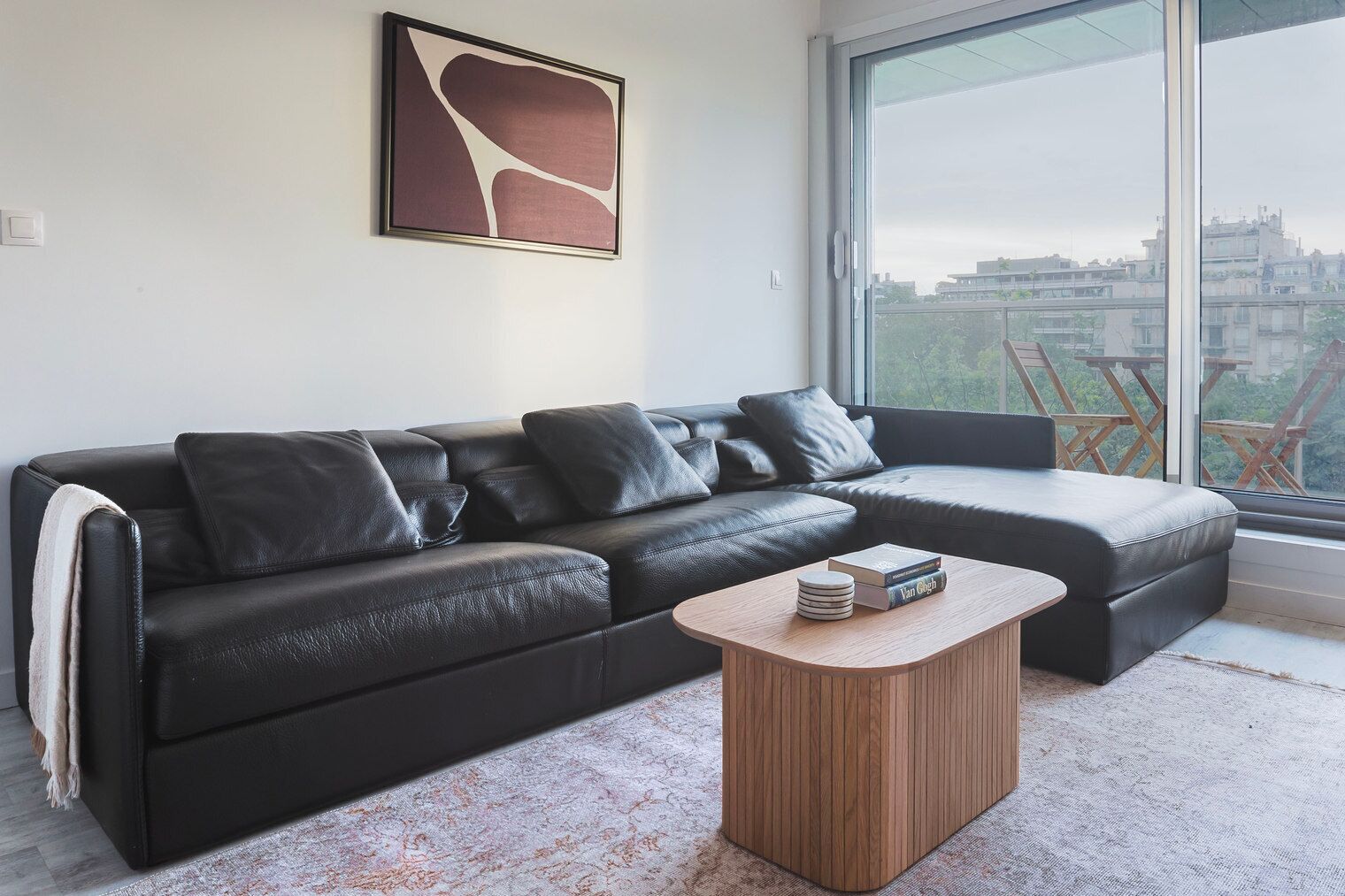 A modern living room with a black leather sofa, a wooden coffee table, and a large window offering a city view.