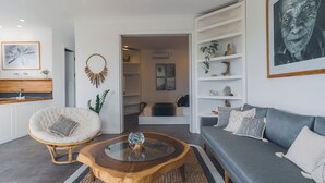 A modern living room with a grey sofa, a round wooden coffee table, a white armchair, and a glass-top coffee table. There is a bookshelf with decorative items and a framed picture on the wall. - Angel Bay (Tabanan)