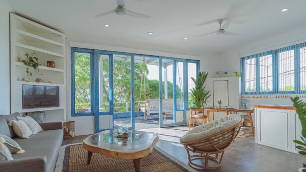 A spacious living area with a sofa, coffee table, and wicker chair. A television is mounted on the wall, and there is a sliding glass door leading to an outdoor patio. - Angel Bay (Tabanan)