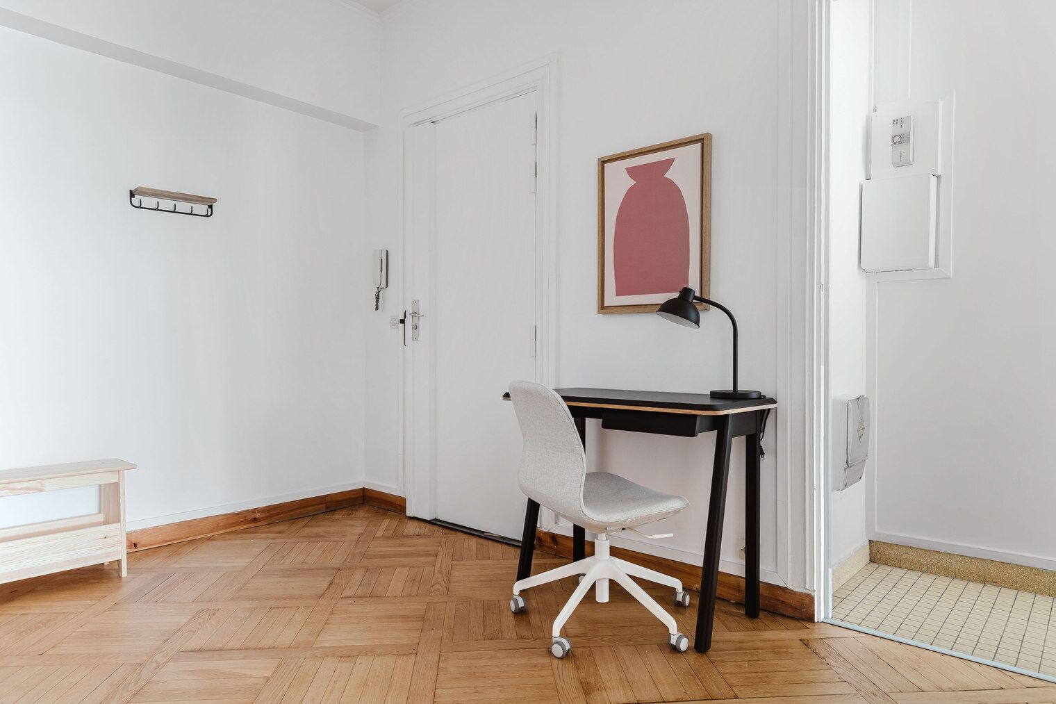 Een kamer met houten vloer, een witte deur, een bureau met stoel, een zwarte bureaulamp en een ingelijst schilderij aan de muur.