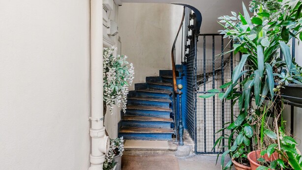Un escalier étroit aux marches bleues menant à un palier, bordé de murs blancs et d’une rambarde en métal noir.