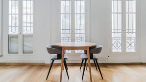 Une pièce avec une table ronde en bois et deux chaises grises à revêtement rembourré. Il y a trois fenêtres à cadre blanc, dotées de grilles en métal, et le sol est en bois.