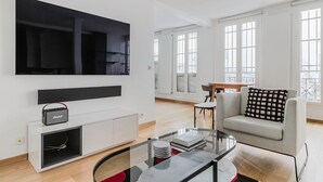 Un salon moderne doté d’un téléviseur à écran plat, d’un petit haut-parleur, d’une table basse en verre, d’un canapé gris et d’un balcon avec de grandes fenêtres.