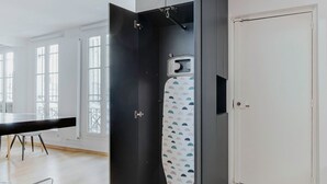 Une pièce avec une porte noire, une planche à repasser blanche ornée d’un motif, une table noire avec une chaise, et une porte blanche.