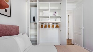 A bedroom with a bed, a closet with shelves, and hanging clothes. - Blueground | Pasteur - Montparnasse, furnished (Paris)