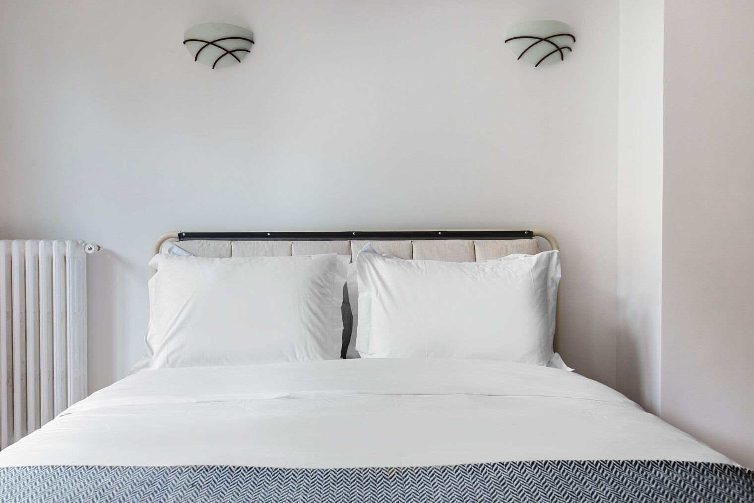 Una cama con sábanas blancas, cabecera y dos lámparas fijadas a la pared.
