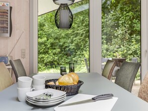 Een eettafel met borden, bekers en een broodmand, voor een raam met uitzicht op groen.