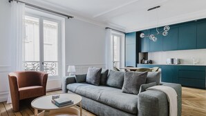 En moderne stue med en grå sofa, en brun lænestol og et rundt træbord. Et køkken med turkise skabe og en hængende lampe er synligt i baggrunden.