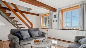 A cozy living room with a grey sofa set, a small round table with a basket of croissants, a wooden staircase, and a large window with curtains. - "Hakon" - 40m from the sea by Interhome (Nexø)