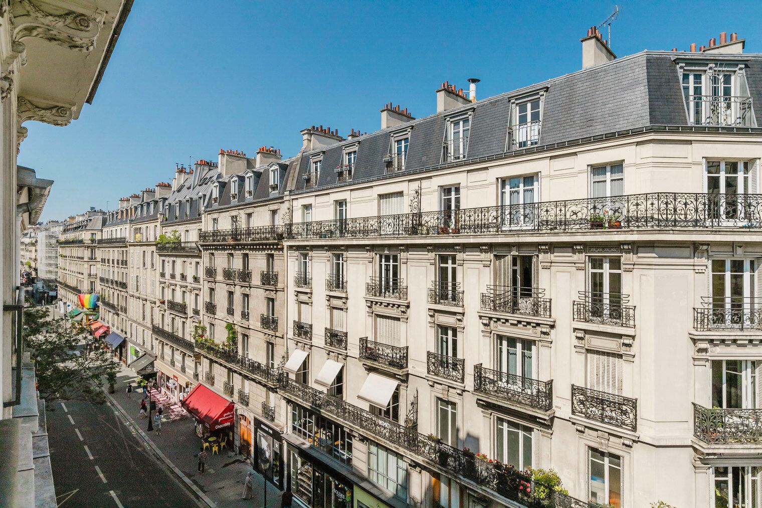 A row of classic European-style buildings with uniform balconies and windows, a clear sky, and a street with shops and pedestrians.