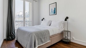 Ein Schlafzimmer mit einem Bett, einem Nachttisch, einer Lampe, einem gerahmten Bild und einem Fenster mit Blick auf Gebäude.