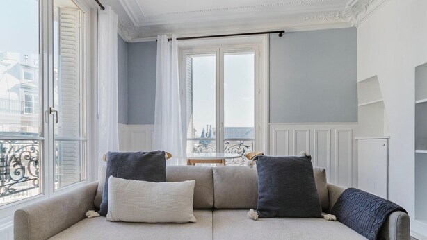 Ein helles Sofa mit drei Kissen in einem Raum mit großem Fenster, weißen Wänden und einem Balkon mit Geländer.
