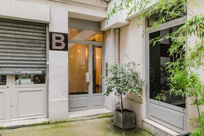 Entrada de un edificio con una persiana cerrada, un letrero con la letra 'B' y una planta en maceta.